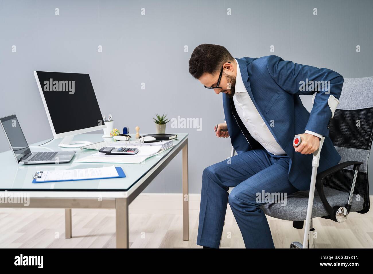 Man With Broken Leg Using Crutches To Get Up From Chair In Office Stock