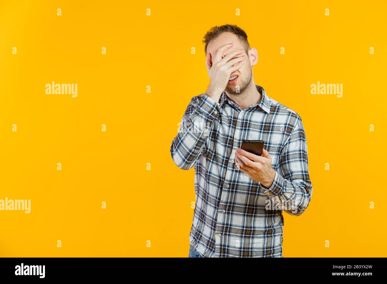 Portrait of tired male student isolated on yellow using his smartphone. Facepalm. Stock Photo