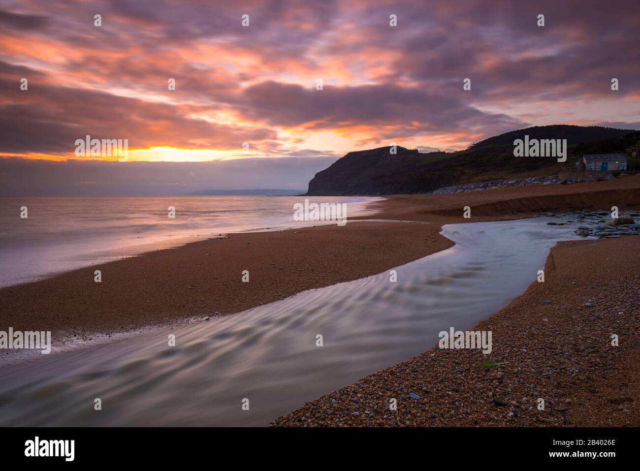Seatown, Dorset, UK.  5th March 2020.  UK Weather.  The clearing clouds glow orange at sunset at Seatown in Dorset after a day of heavy rain with the River Winniford flowing across the beach and in the distance is the distinctive shaped cliff of Golden Cap which is the highest cliff on the South Coast of England.  Picture Credit: Graham Hunt/Alamy Live News Stock Photo