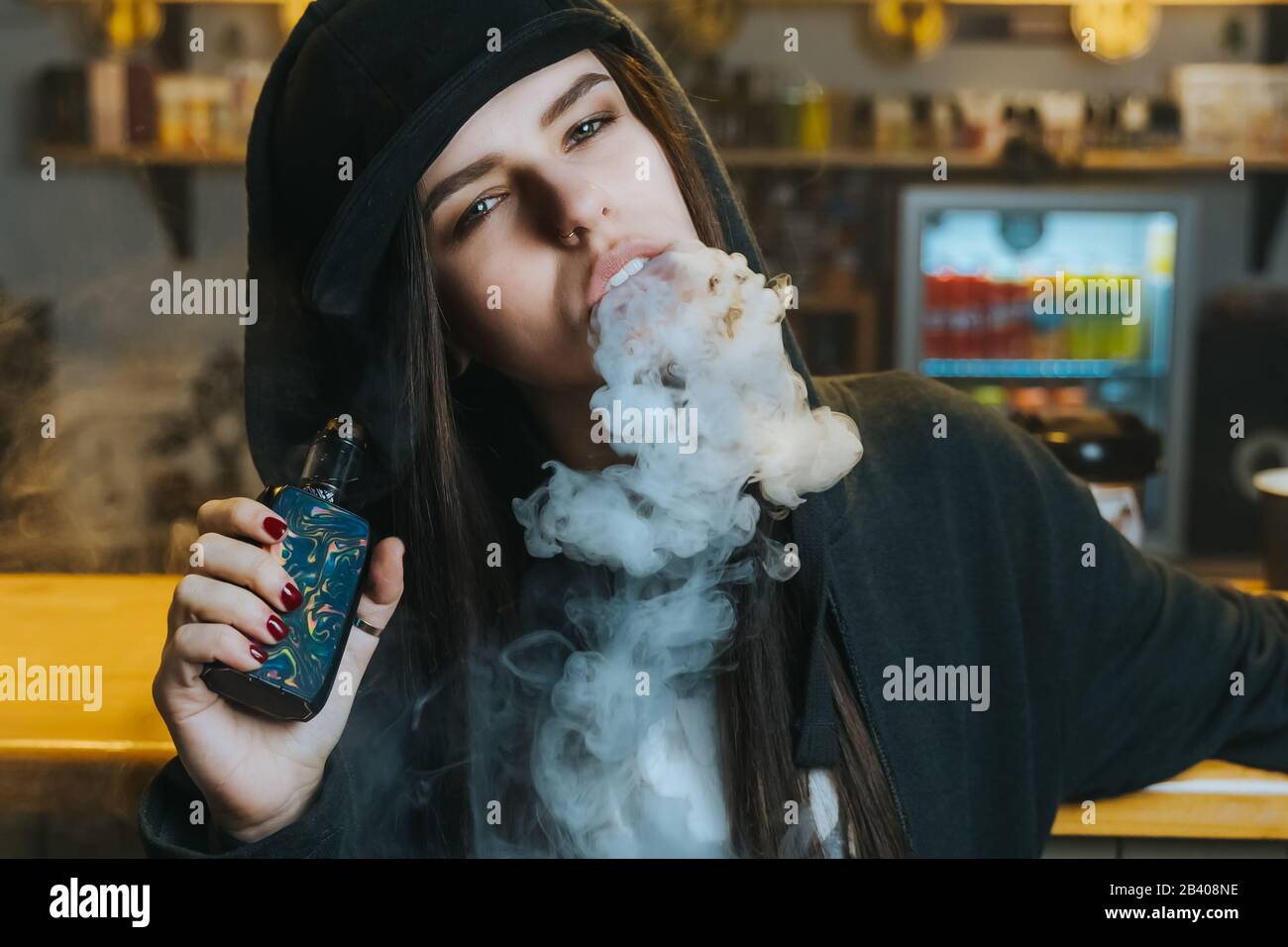 Young pretty woman in cap smoke an electronic cigarette at the vape shop. Hip-hop style. Closeup. Stock Photo