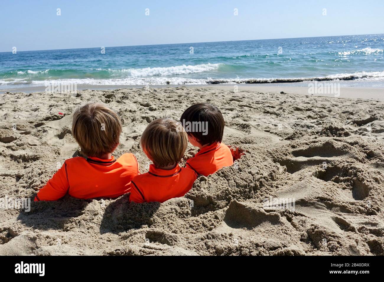 Three cousins sit buried in the sand while looking out at the ocean. Stock Photo