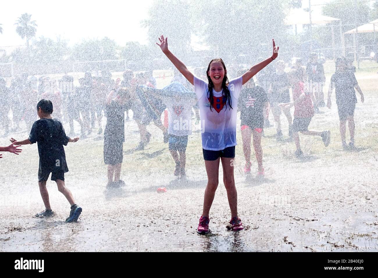 A girl throws her hands in the air and smiles while being drenched by a fire hose during a fun school field day. Stock Photo