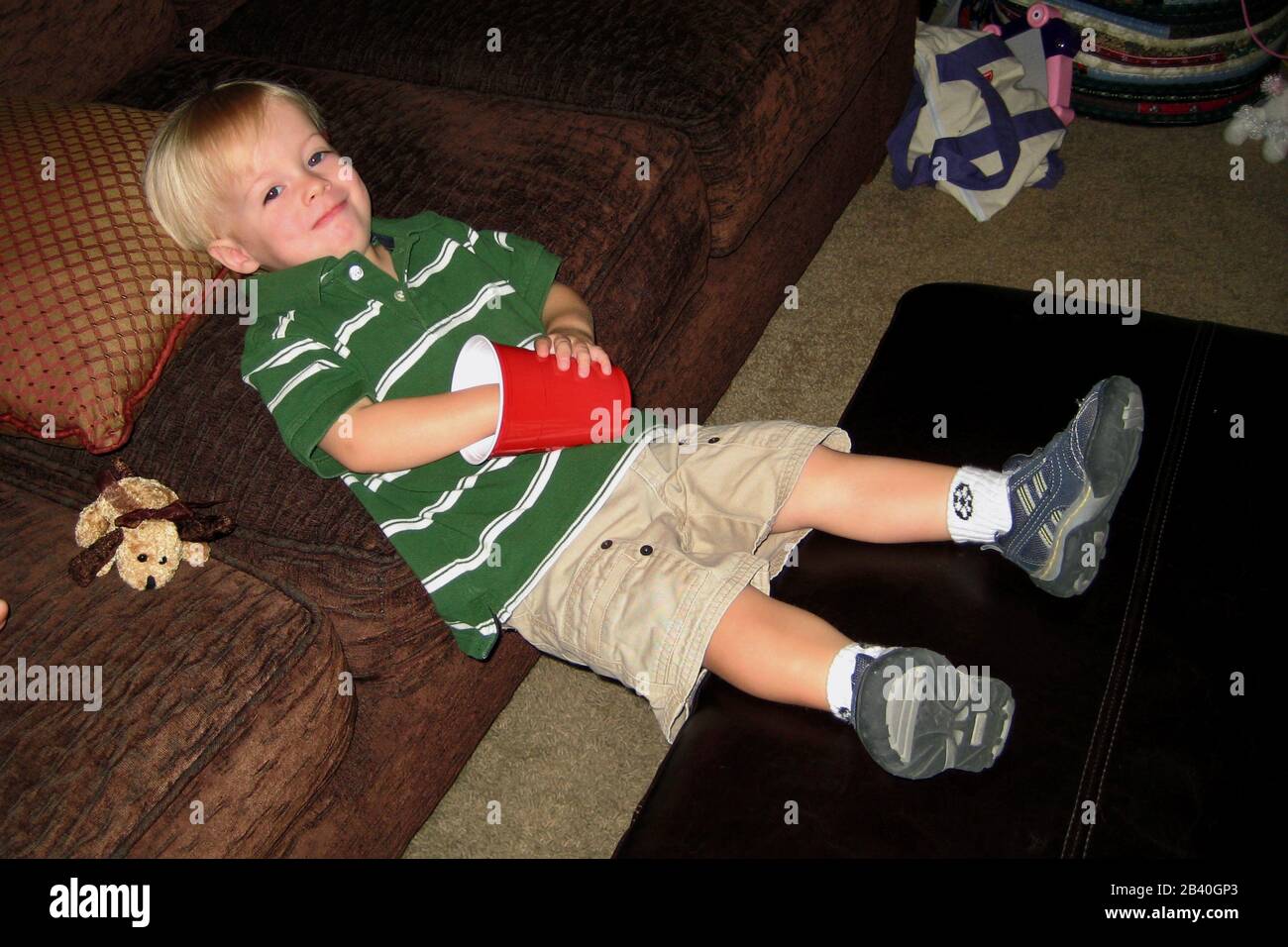 A little boy stretches out on the edge of a couch while eating a snack from a red cup. Stock Photo