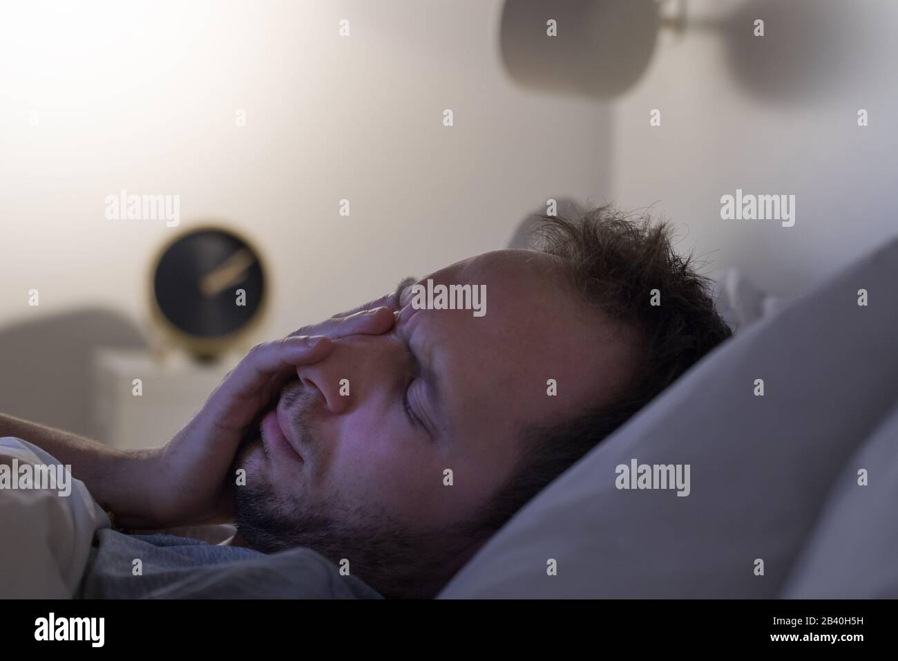 Insomnia, bad dreams concept. Man can't sleep after party, lying on bed at night, alarm clock on bedside table. Sleep/wake cycle disorders Stock Photo
