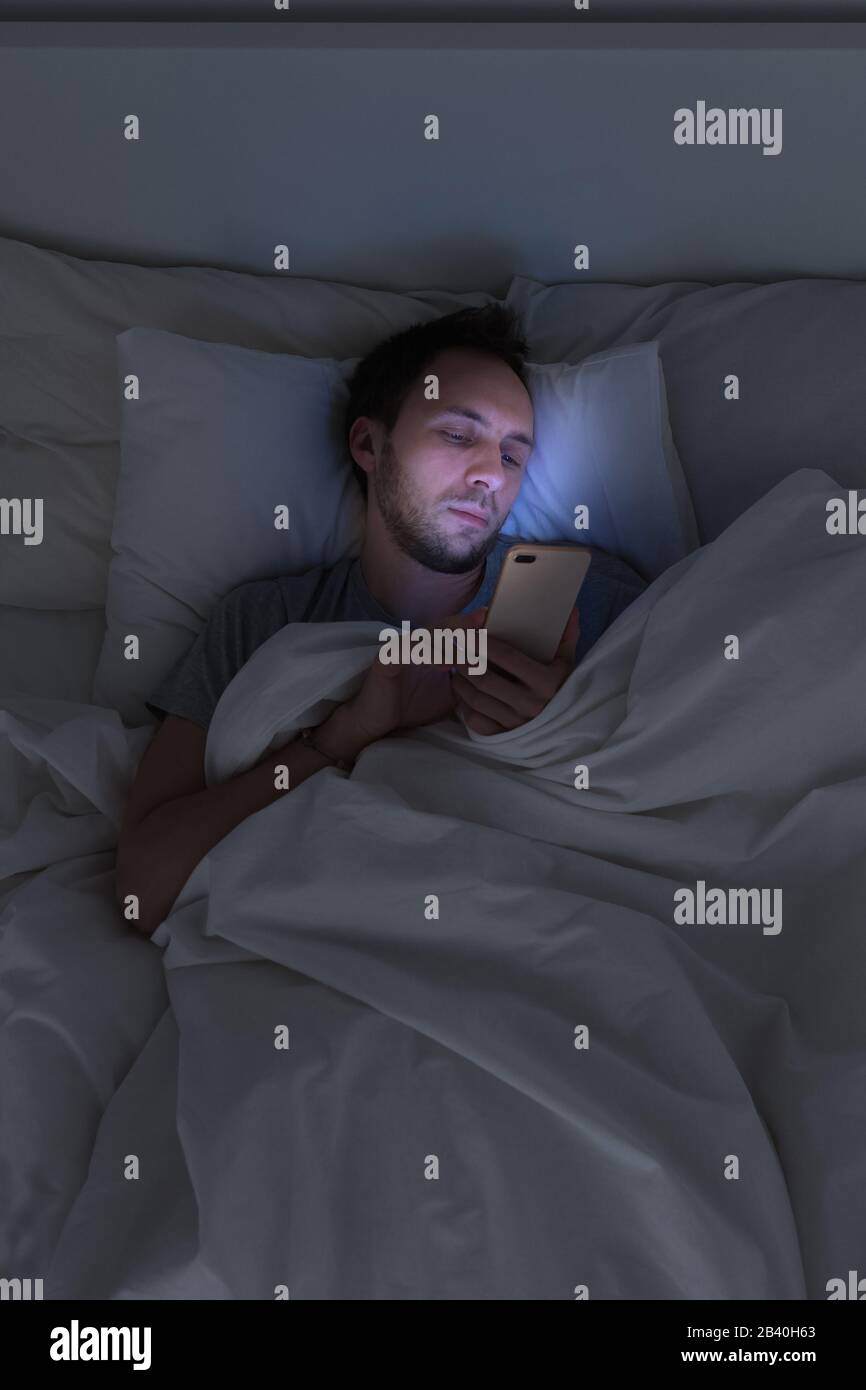 Mobile addiction, nomophobia, insomnia, sleep disorder concept. Bored man chatting and surfing on internet with smart phone late at night, lying on be Stock Photo
