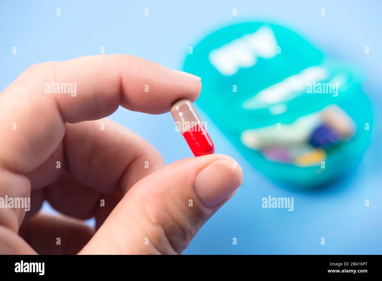 Colorful capsule in patient's hand with daily pill dispenser on blue background. Stock Photo