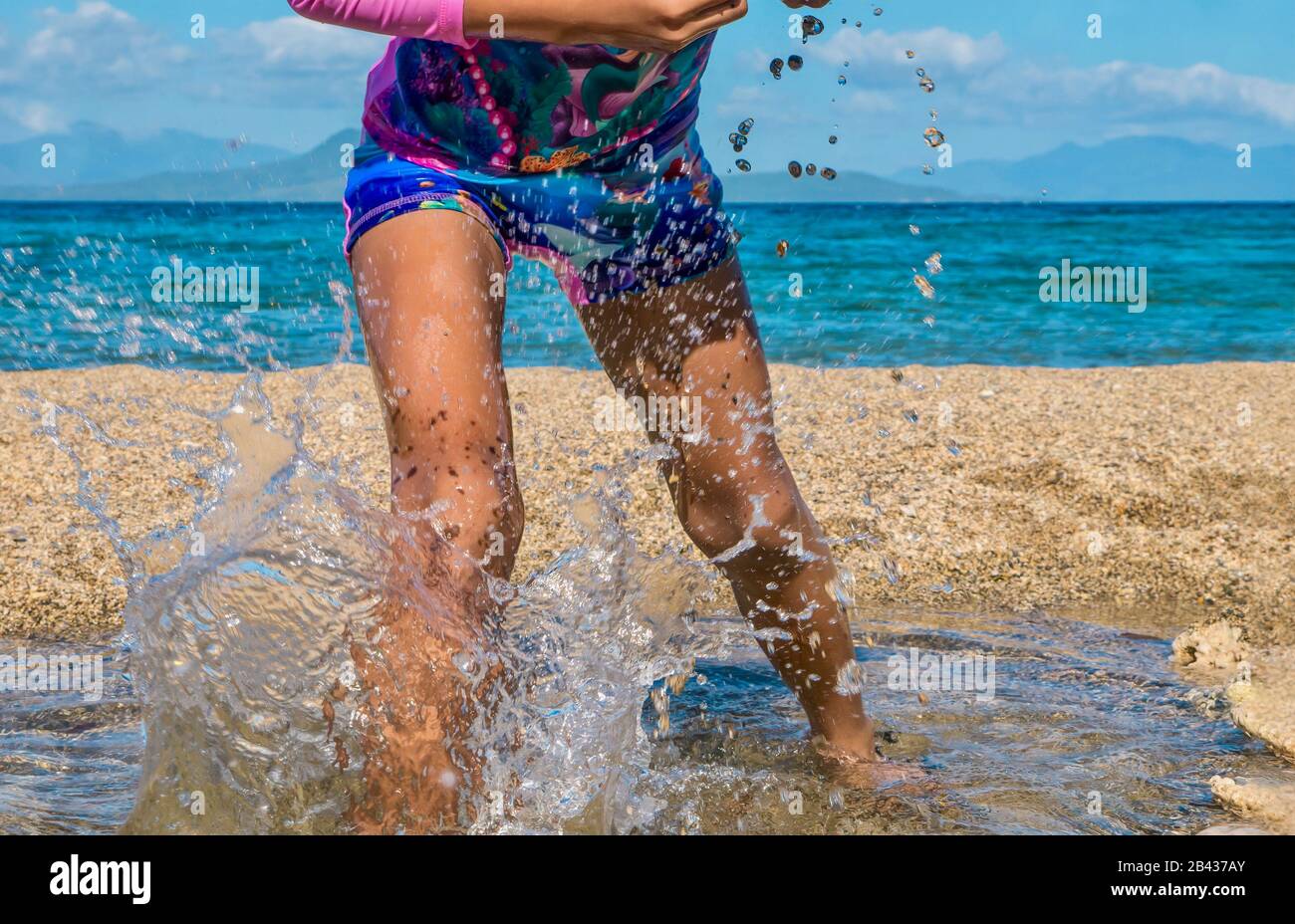 Action shot of an unidentifiable girl having fun, playfully splashing in the water during a summer holiday on an island beach. Legs and waist visible. Stock Photo