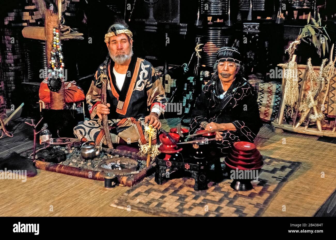 An elderly Ainu couple pose in traditional dress with souvenirs they sell in a native home that is open to tourists on the island of Hokkaido in northern Japan. The 70-year-old bearded man sits next to his 84-year-old aunt with a tattoo around her mouth that resembles a mustache. The painful tradition of rubbing soot into cuts in the skin during childhood to ward off evil spirits and to indicate a girl was ready for marriage has since been outlawed by the Japanese government. The woman was one of only 300 pure-blooded Ainu still living when this historical photograph was taken in 1962. Stock Photo