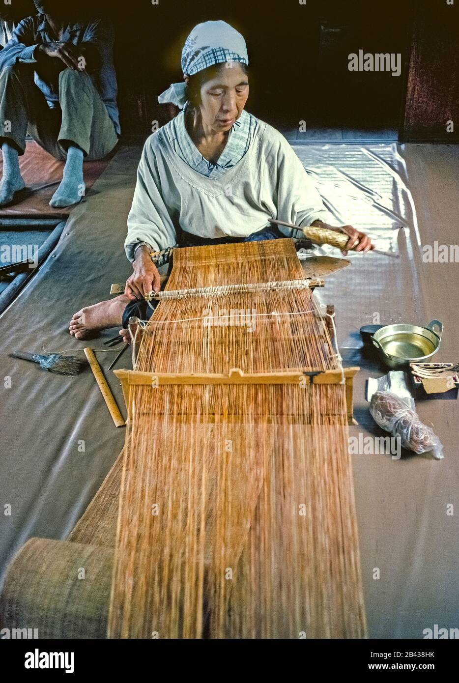 An Ainu woman uses the tedious traditional way to weave matting while sitting on the floor of her native home on the island of Hokkaido in northern Japan. The material is then made into placemats and tablecloths to sell as souvenirs in special Ainu villages that give Japanese tourists a glimpse of bygone aboriginal life. Only 300 pure-blooded Ainu (pronounced I-noo) were still living when this historical photograph was taken in 1962. The Ainu were officially recognized as indigenous people of Japan in 2008. Stock Photo