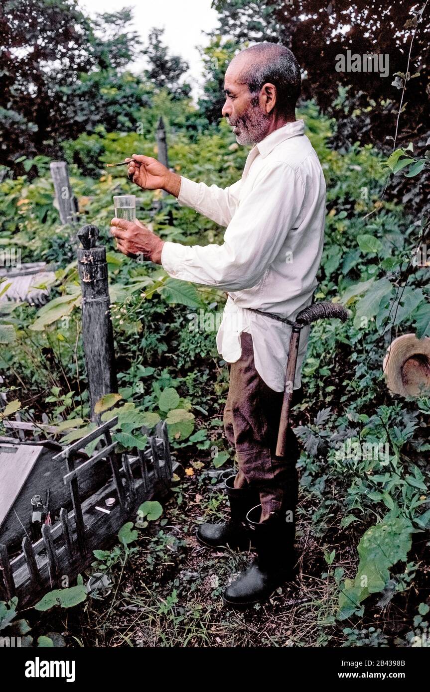 An elderly Ainu man sprinkles shochu (Japanese liquor) on the overgrown graves of ancestors in an ancient Ainu village graveyard on the island of Hokkaido in northern Japan. The beverage is offered with prayers to the dead as a gesture of goodwill and friendship. Ainu grave markers are wooden posts with carvings at the top representing the hole in a needle to indicate a woman is buried there, or a spear point to represent the burial site of a man. The Ainu (pronounced I-noo) were officially recognized as indigenous people of Japan in 2008. This historical photograph was taken in 1962. Stock Photo