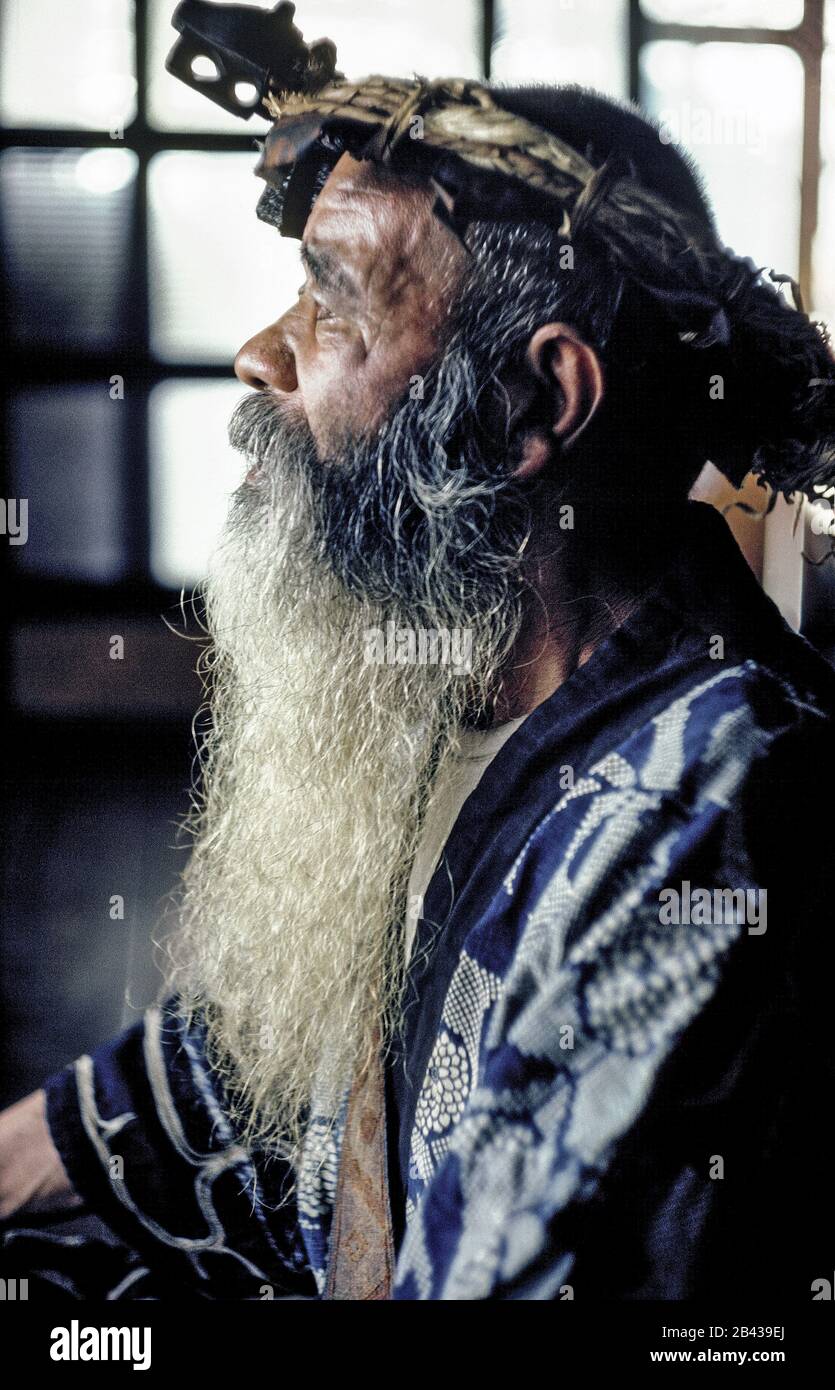 An Elderly Ainu Man With A Long Beard And Wearing A Crown Of Wood Fibers Poses In Traditional Dress Inside His Rural Home On The Island Of Hokkaido In Northern Japan His