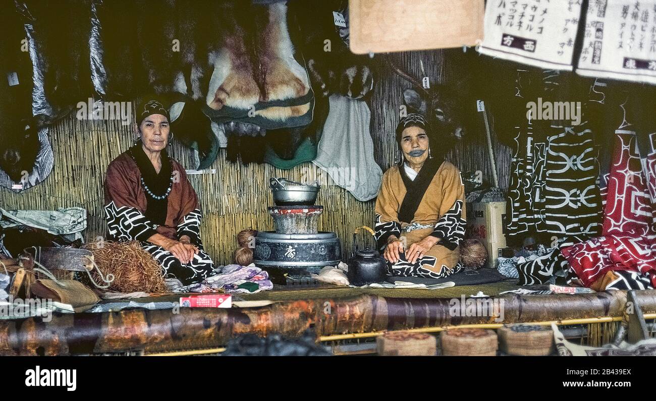 Two elderly Ainu women in traditional dress sell souvenirs in an Ainu village on the island of Hokkaido in northern Japan. A tattoo around her mouth that resembles a mustache is evident on the face of one woman. The painful tradition of rubbing soot into cuts in the skin during the childhood of Ainu girls to ward off evil spirits has since been outlawed by the Japanese government. The women were two of only 300 pure-blooded Ainu (pronounced I-noo) still living when this historical photograph was taken in 1962. The Ainu were officially recognized as indigenous people of Japan in 2008. Stock Photo