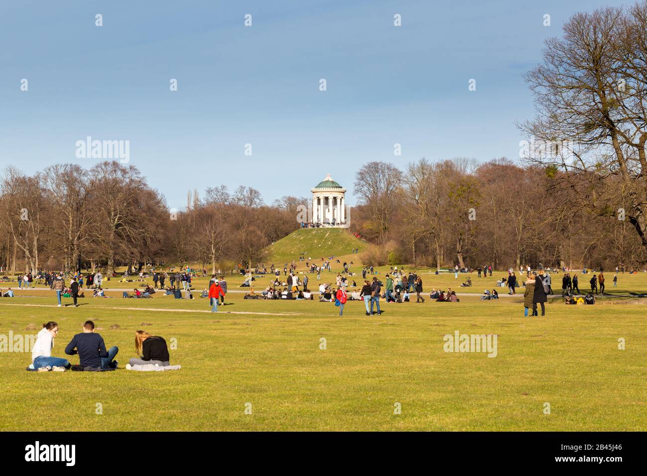 View on the so-called Monopteros at the English Garden (Englischer Garten). In the foreground people walking by & sitting in the grass. Popular park. Stock Photo