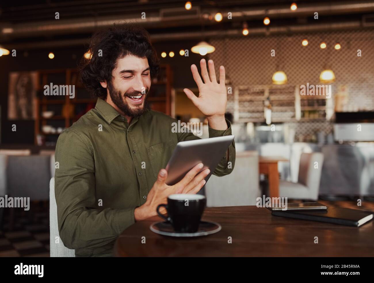 Casual bearded man waving his hand while talking through video-chat in smartphone while relaxing in cafe Stock Photo