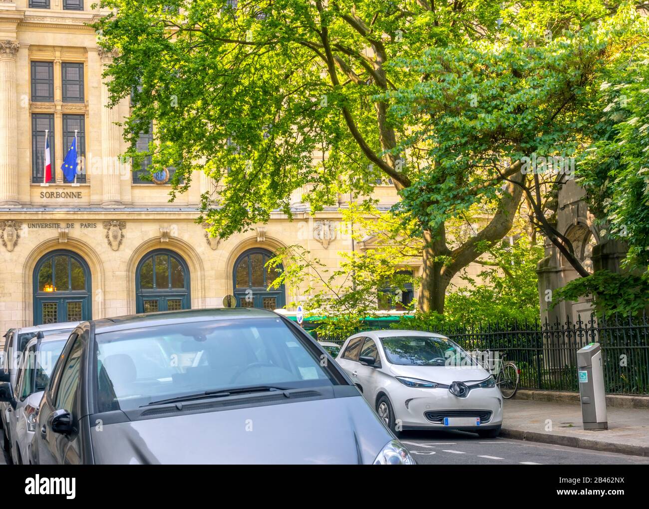 France. Summer sunny day in Paris. Green garden near the University of Sorbonne. Several cars are parked on a narrow street. Electric car connected to Stock Photo