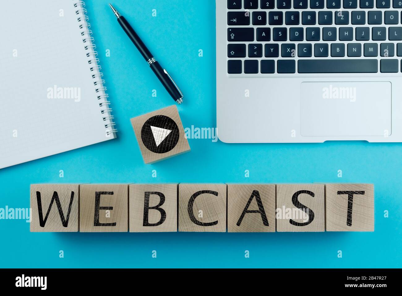 word WEBCAST and play button symbol on wood cubes against blue background with laptop and notepad, webinar and web broadcast concept Stock Photo