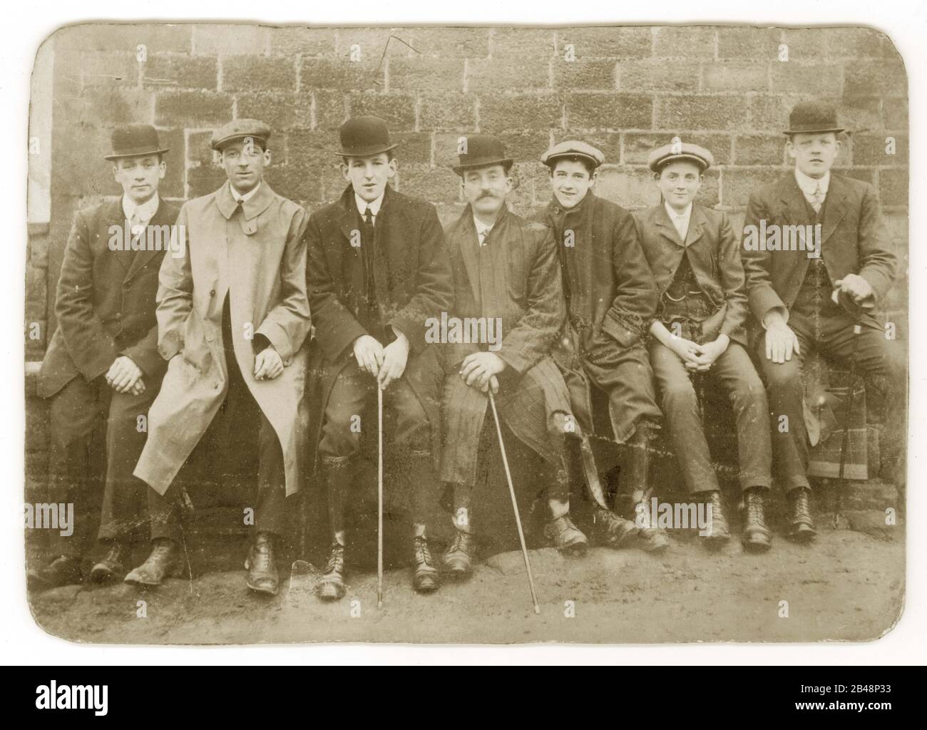 Early 1900's Edwardian cabinet card of group of seven young men and boys wearing flat caps and bowler hats, some holding walking sticks or canes, rounded collars, sitting together resting after a walk on a bench, U.K. Circa 1905 Stock Photo