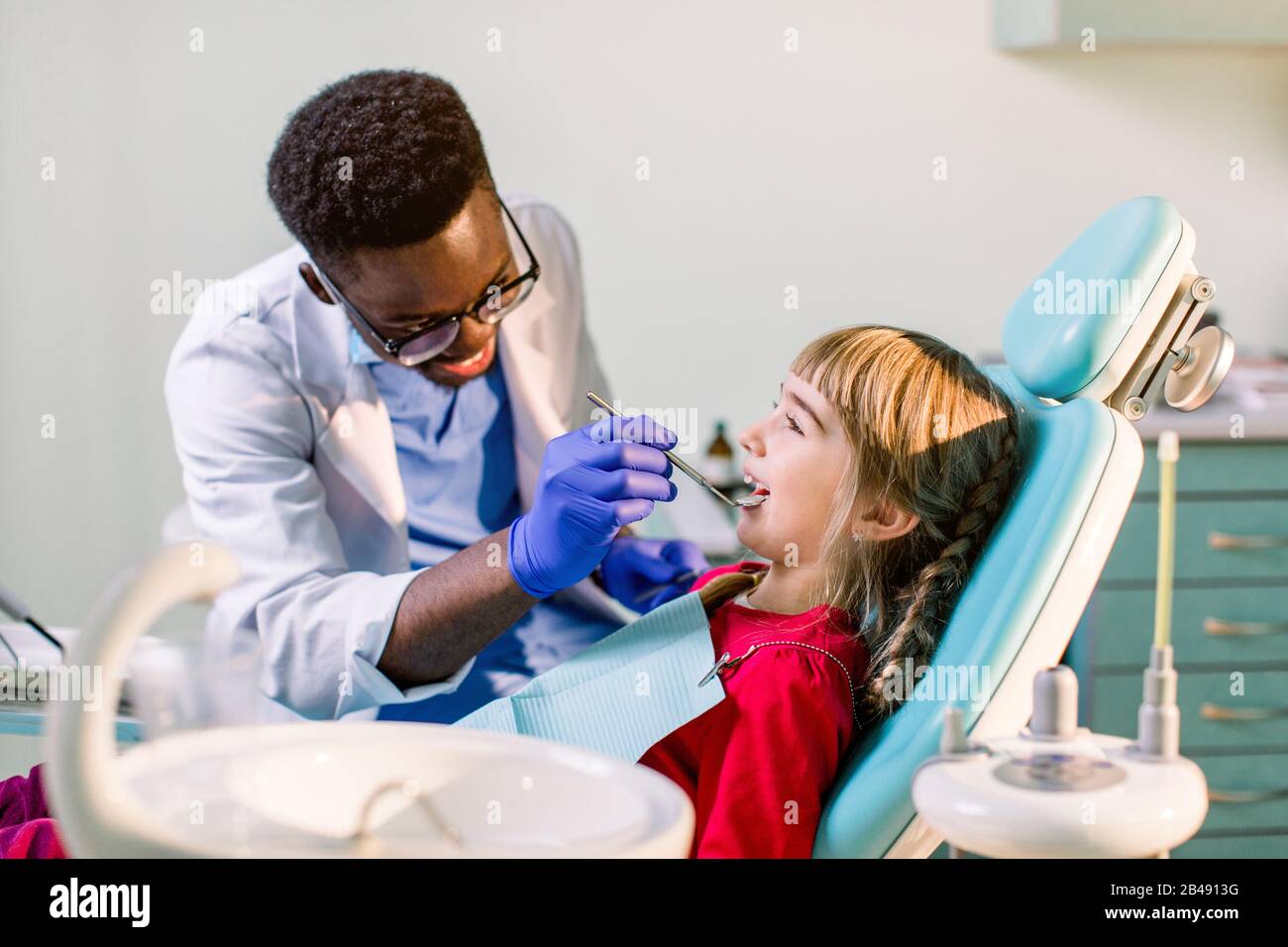 African-American ethnic black male dentist in blue latex gloves checking  condition of teeth of little smiling girl. baby girl in blue dental chair  in Stock Photo - Alamy