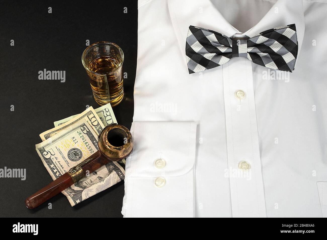 white dress shirt and black and white checkered bow tie with shot of whiskey and smoking pipe on American money Stock Photo