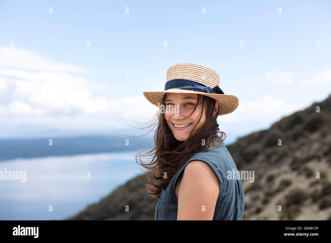 Frau im Leinenkleid und mit Sonnenhut steht an Küste Kroatiens und schaut lächelnd zum Meer Stock Photo