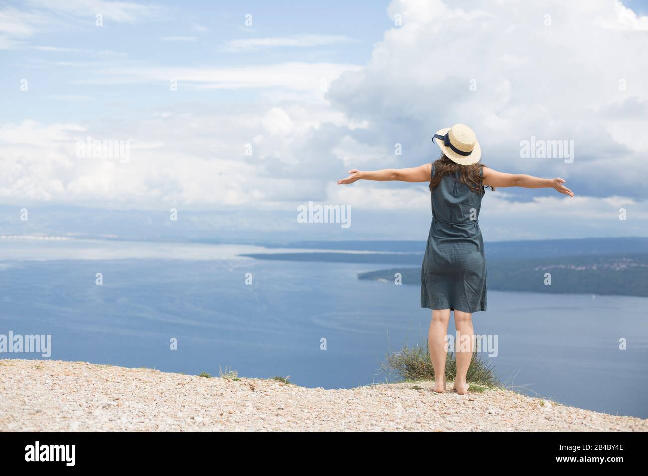 Woman in linen dress and sun hat is standing on coast of Croatia, spreads arms and looks to the sea Stock Photo