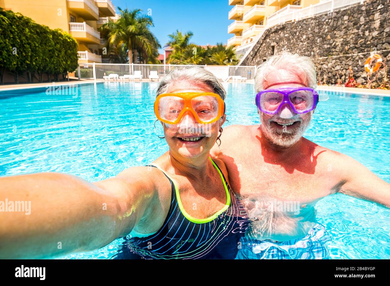 Happy cheerful people old senior man and woman have fun together in the summer swimming pool activity taking selfie pictures with scuba masks on the face for funny outdoor leisure activity in hotel holiday vacation Stock Photo