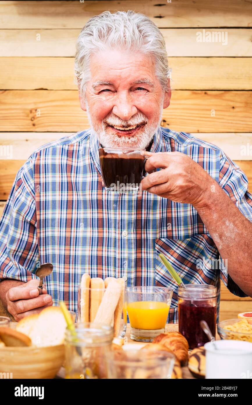 Cheerful and funny active nice senior man drinking hot chocolate with beard and moustache dirty - happy retired people enjoying breakfast - wooden style wall in background - table full of food Stock Photo