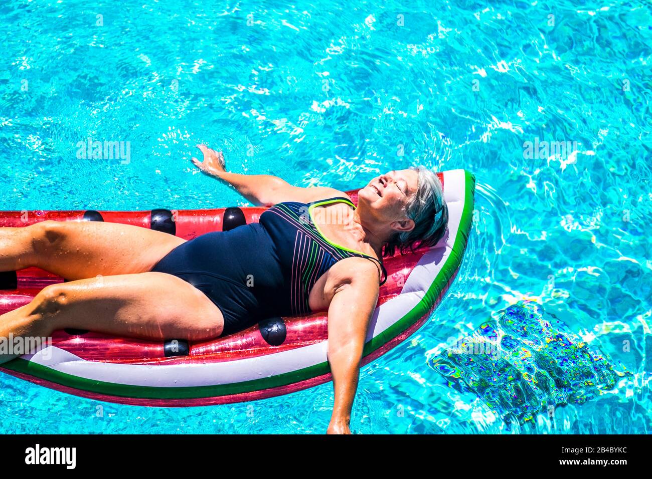 Happy beautiful aged senior lady enjoy the fresh water on a watermelon coloured lilo inflatable mattress on the blue water of swimming pool smiling and relaxing in a sunny day of summer Stock Photo