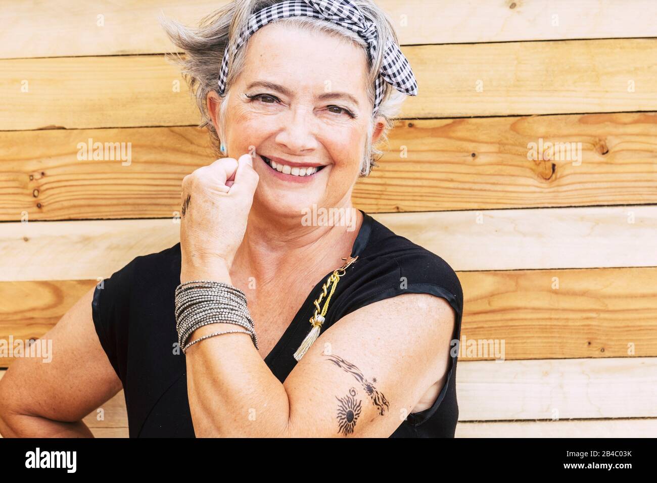 Rock and alternative cheerful senior caucasian woman show her tatoo on the arm smiilng at the camera - no limit age to have fun and enjoy lifestyle for old people - wooden background Stock Photo