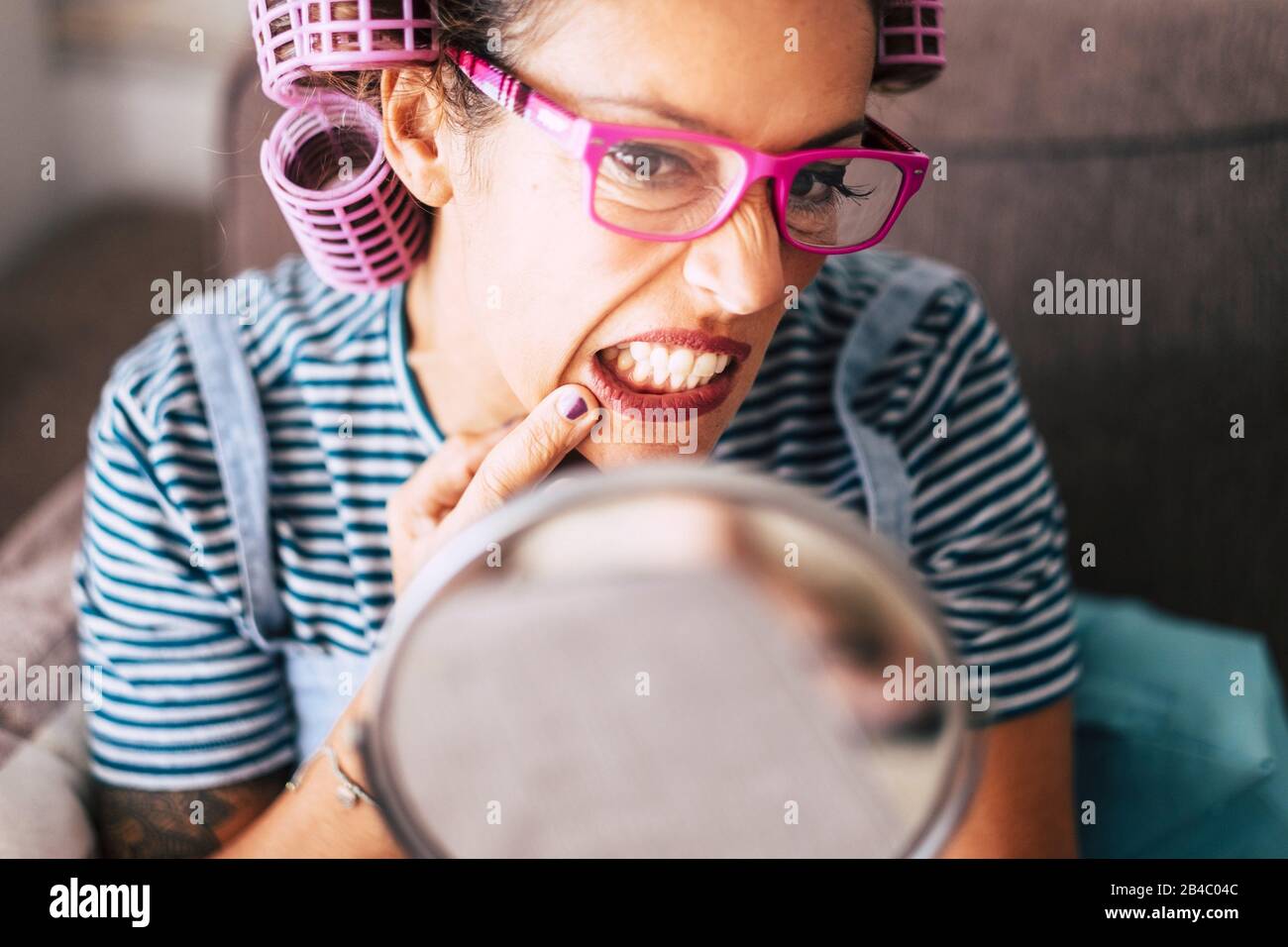 Nice and beautiful young adult woman at home checking skin wrinkles or age imperfections in front of a mirror - curlers on hair and eyeglasses for the adult age - active modern people live healthy with stress Stock Photo