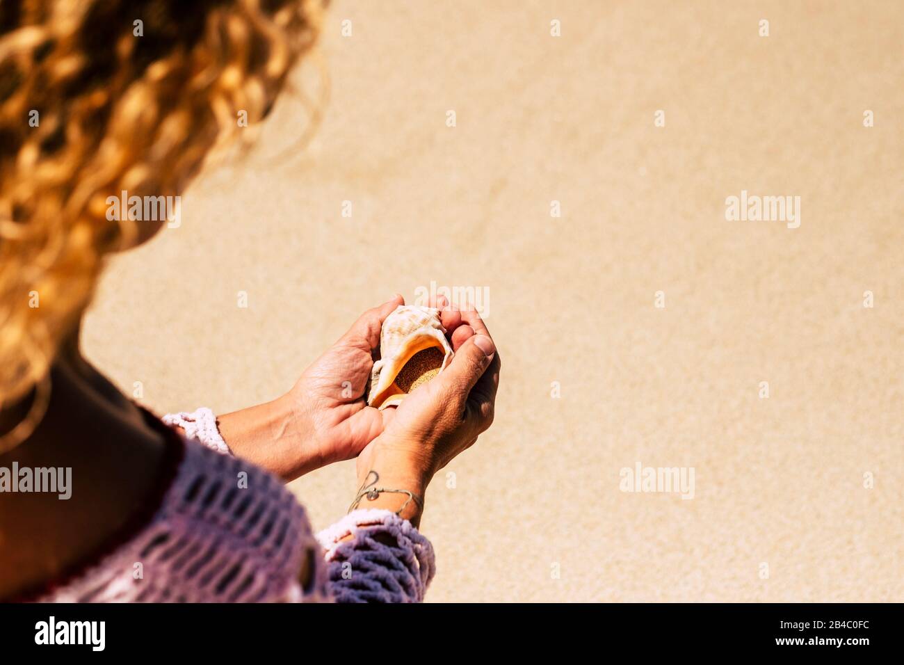 Point of view above shoulder with blonde woman holding a beautiful seashell on hands with sand inside - earth's day concept and respect for nature - beach in background for summer holiday vacation Stock Photo