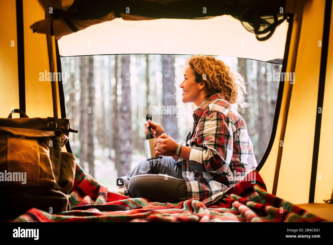Camping with tent and adventure alternative travel vacation concept with cheerful people - beautiful adult blonde smile and enjoy the outdoors nature around in the forest sit down near a backpack and drinking coffee Stock Photo