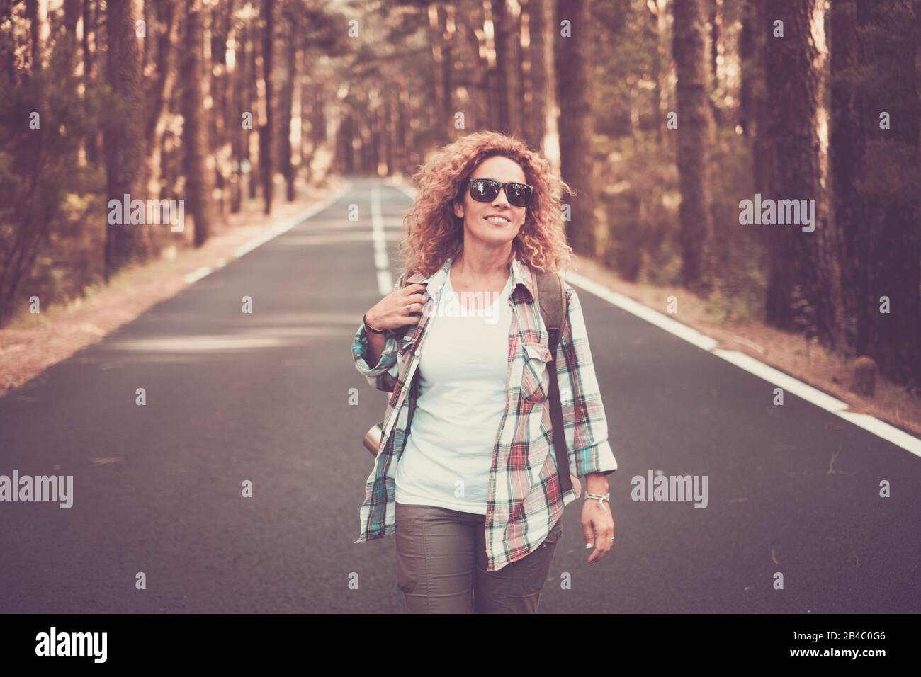 Cheerful happy free woman traveler people walking in the middle of a long road with forest and trees around - scenic travel concept - beautiful caucasian curly female enjoying the outdoor leisure activity Stock Photo