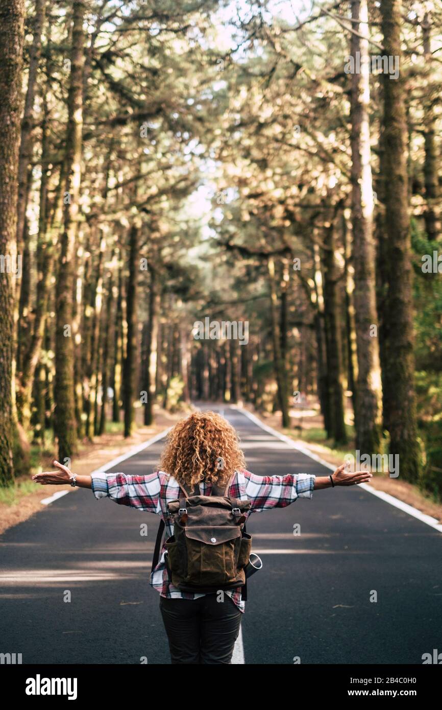 Alternative travel concept with curly hipster woman viewed from back open his arms and feel the freedom of the outdoors nature standing in the middle of a ling road - happiness and wanderlust lifestyle Stock Photo