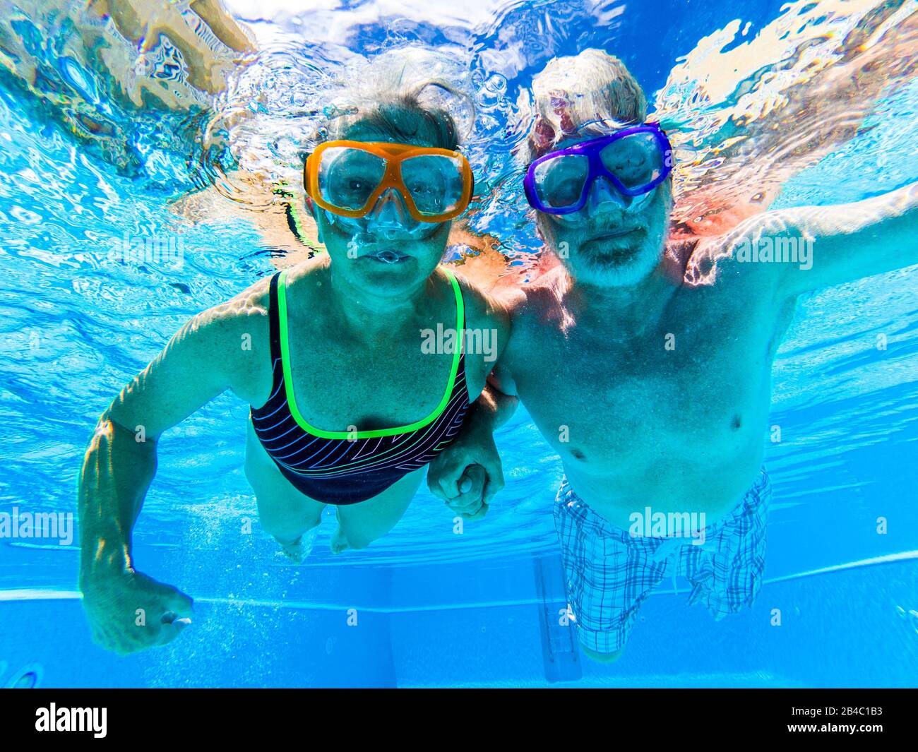 Adult people senior couple have fun swimmin in the pool underwater with coloured funny diving masks - dive concept and active retired man and woman enjoying the lifestyle - blue water and caucasian adults Stock Photo