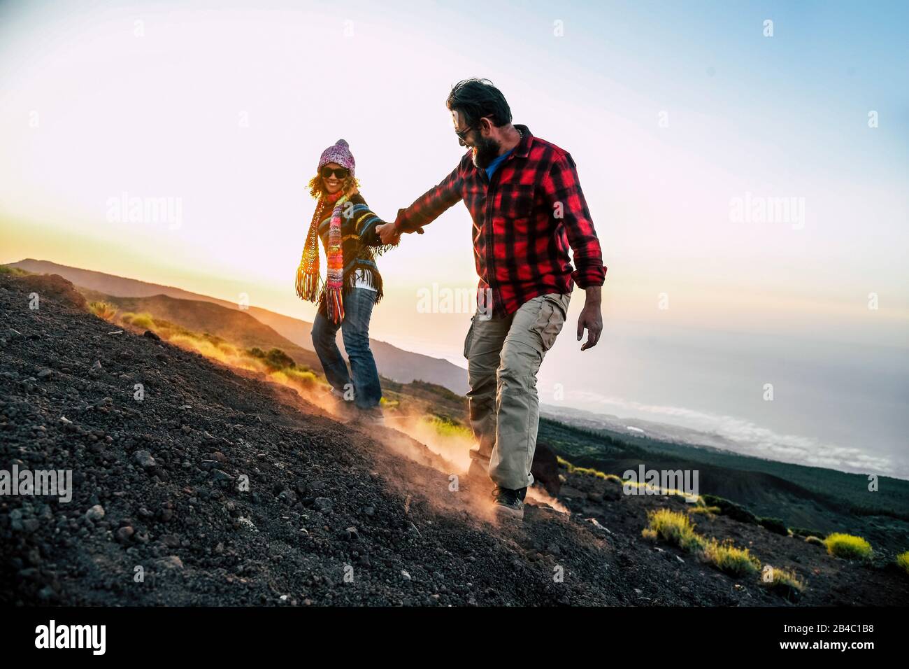 Happy people in outdoor leisure activity at the mountain - trekking active couple holding hand to help and have fun together - beautiful scenic sunset landscape in background - travel and adventure Stock Photo