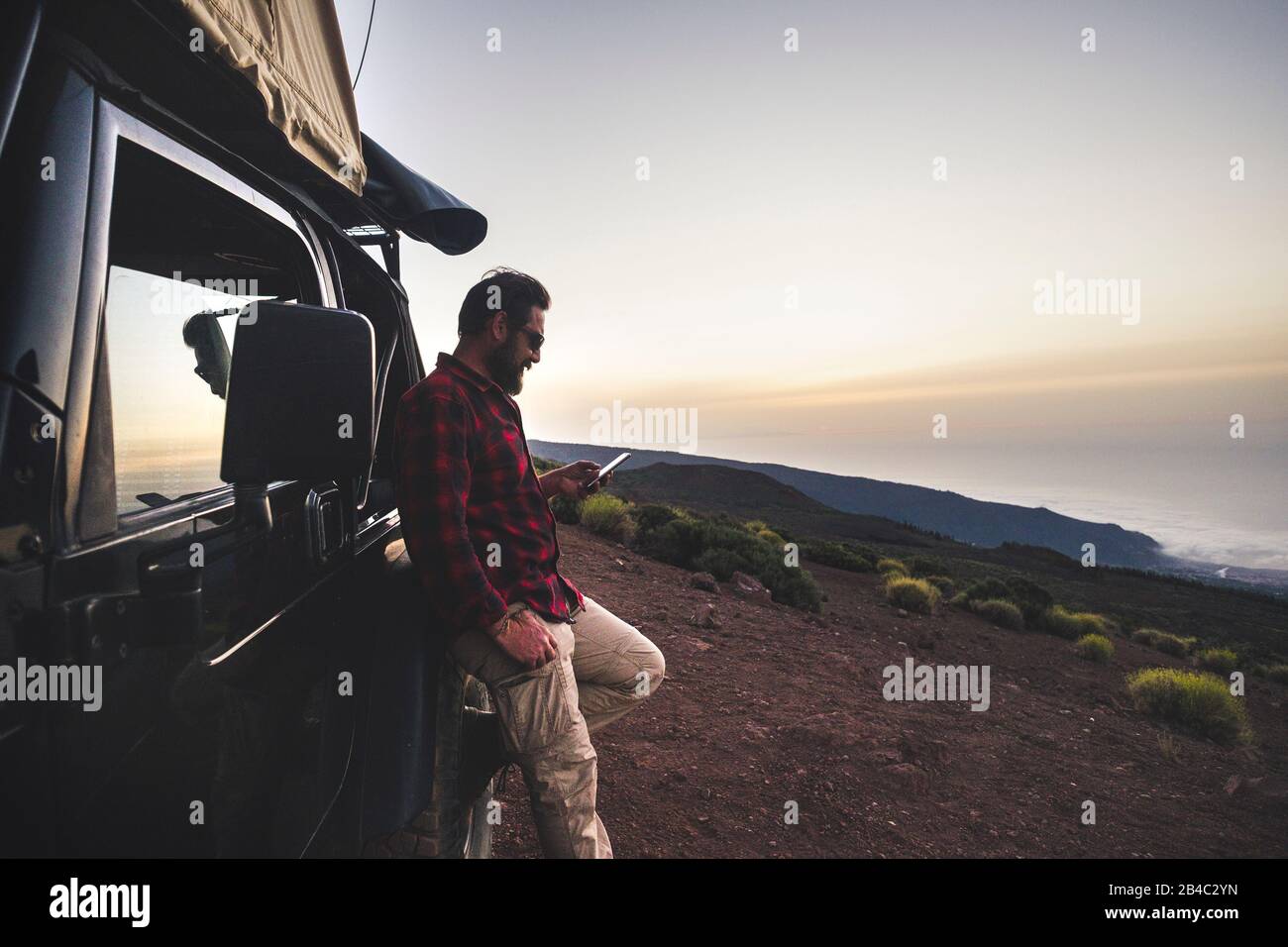 Adventure explorer traveler man use cellular phone with internet connection in wild mountain place during travel excursion with off road black car and tent on the roof - free people concept Stock Photo