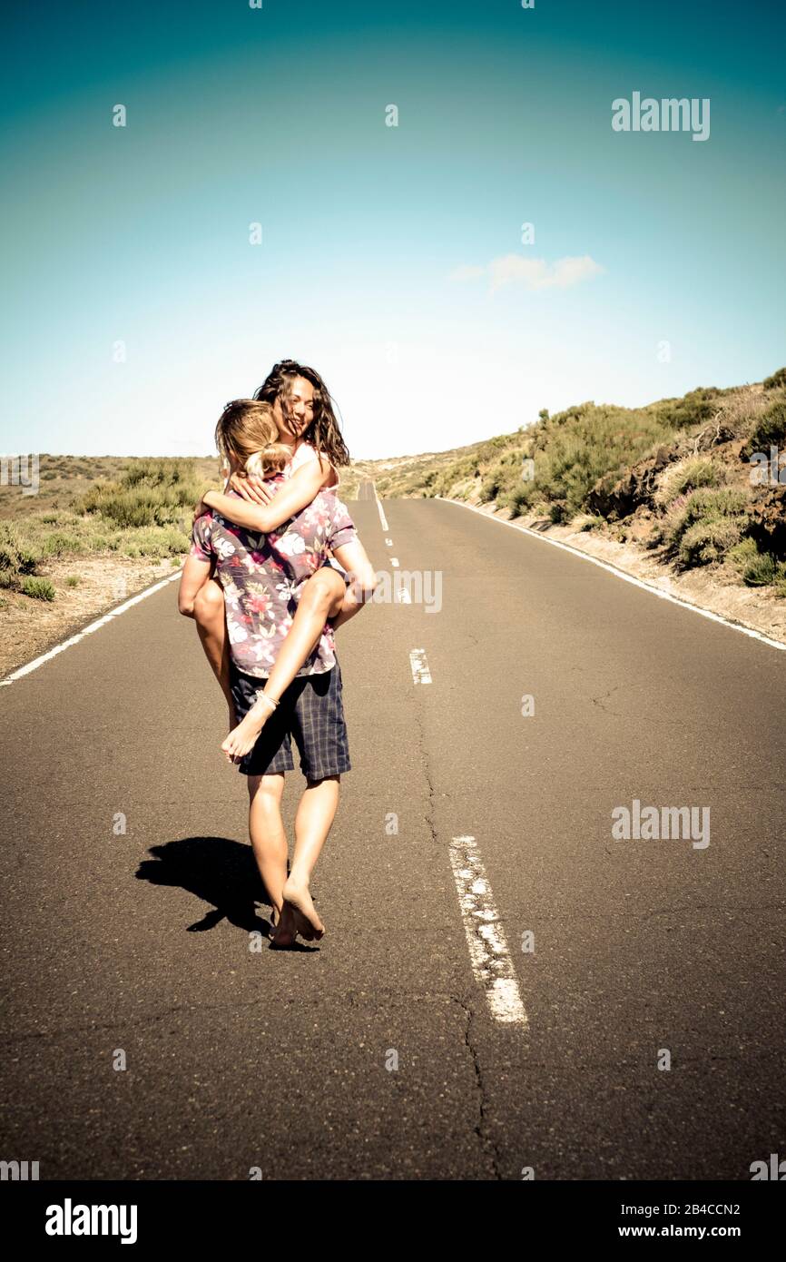 Happy young couple in love walking together on a long straight road - man carry laughing cheerful woman - concept of travel together and have fun Stock Photo