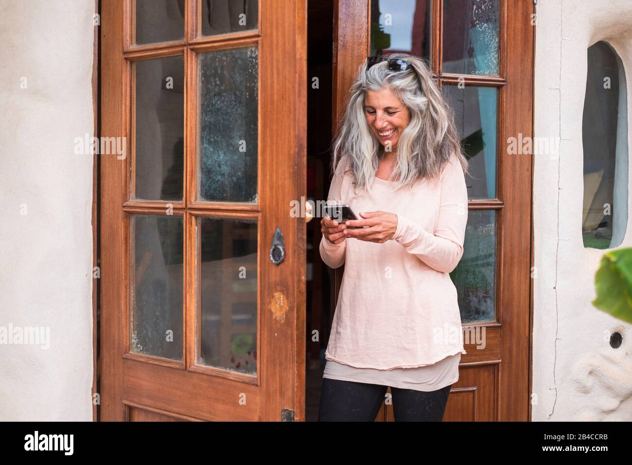 Happy and cheerful adult alternative woman using technology outside home in the garden - long grey hair and smille using a mobile phone out of the door Stock Photo