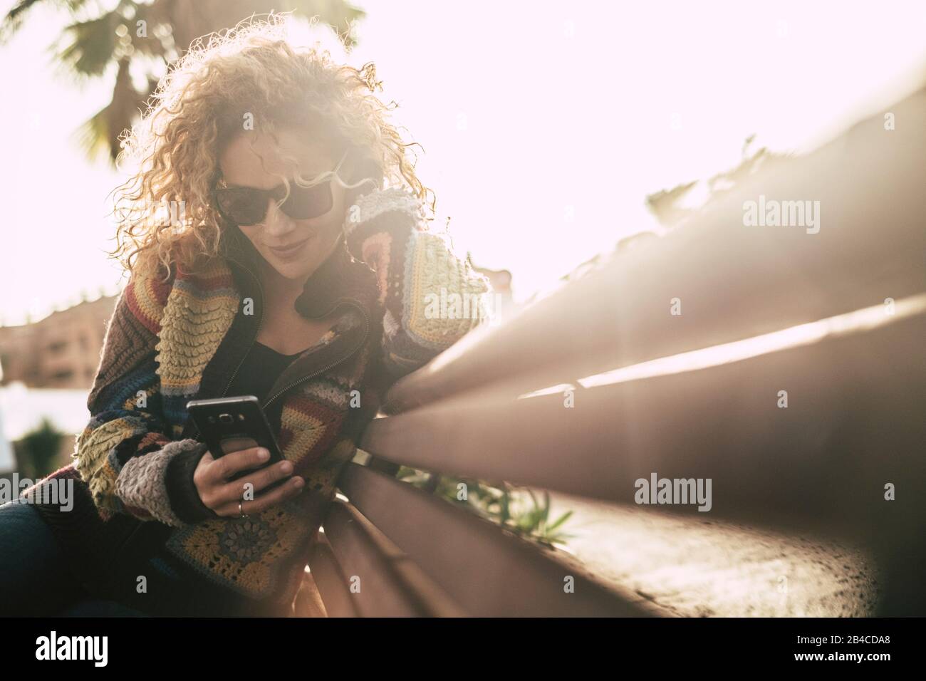 Autumn season colors with beautiful adult attractive blonde curly woman use mobile phone sitting on a beanch in outoor leisure activity - sun in backlight and bright background Stock Photo