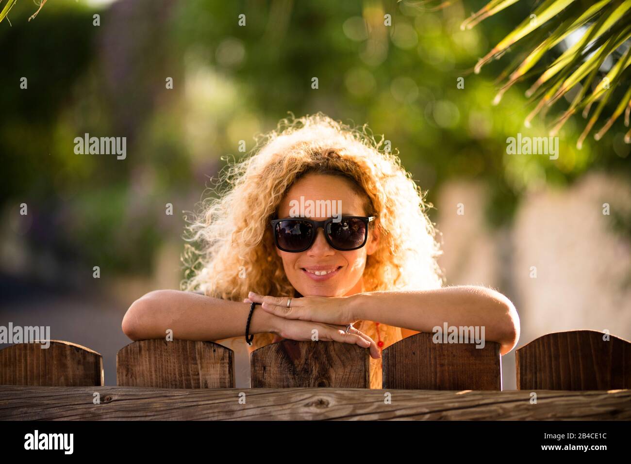 Portrait of beautiful caucasian young blonde curly woman cheerful looking at the camera with sun lights in background and defocused green natural outdoor background - happy middle age lady concept Stock Photo