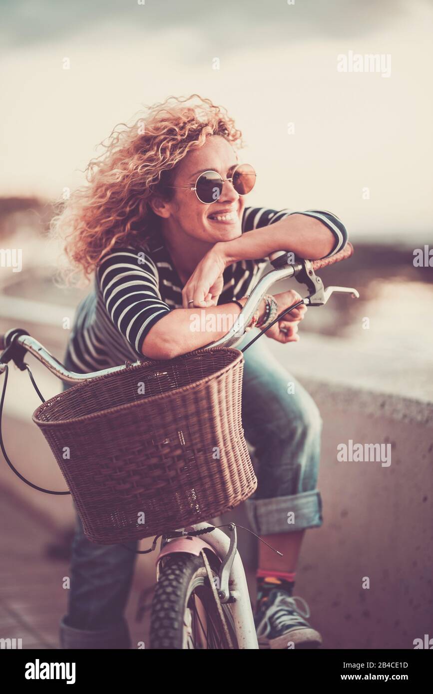 Cheerful trendy young adult caucasian woman sitting on a bike and smiling - beautiful female portrait - concept of outdoor leisure activity and happiness and joyful lifestyle Stock Photo
