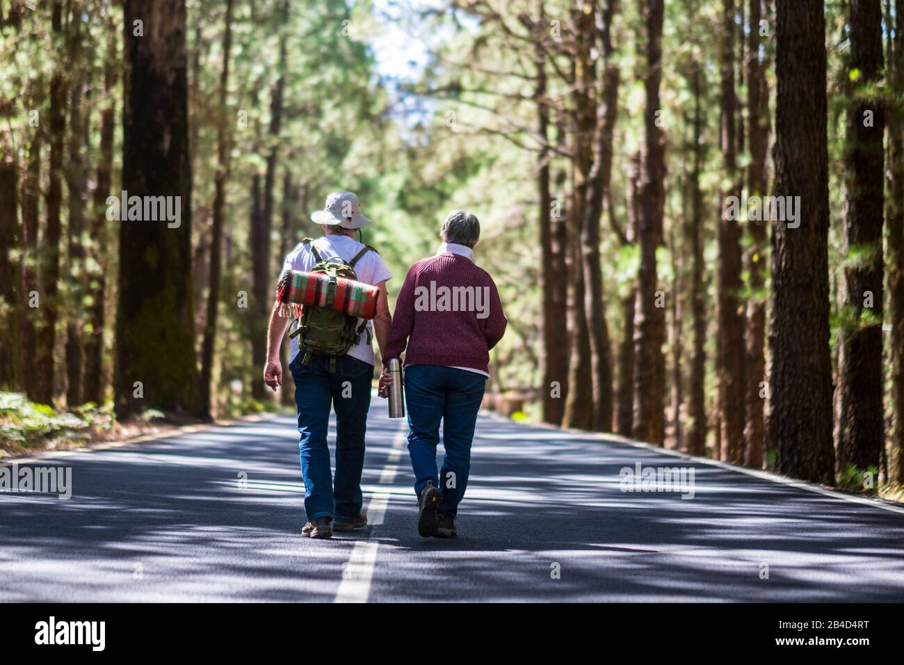 Travel lifestyle for old senior couple walking together in the middle of the road with high trees forest on both sides - lifestyle and togetherness forever life concept - aged people Stock Photo