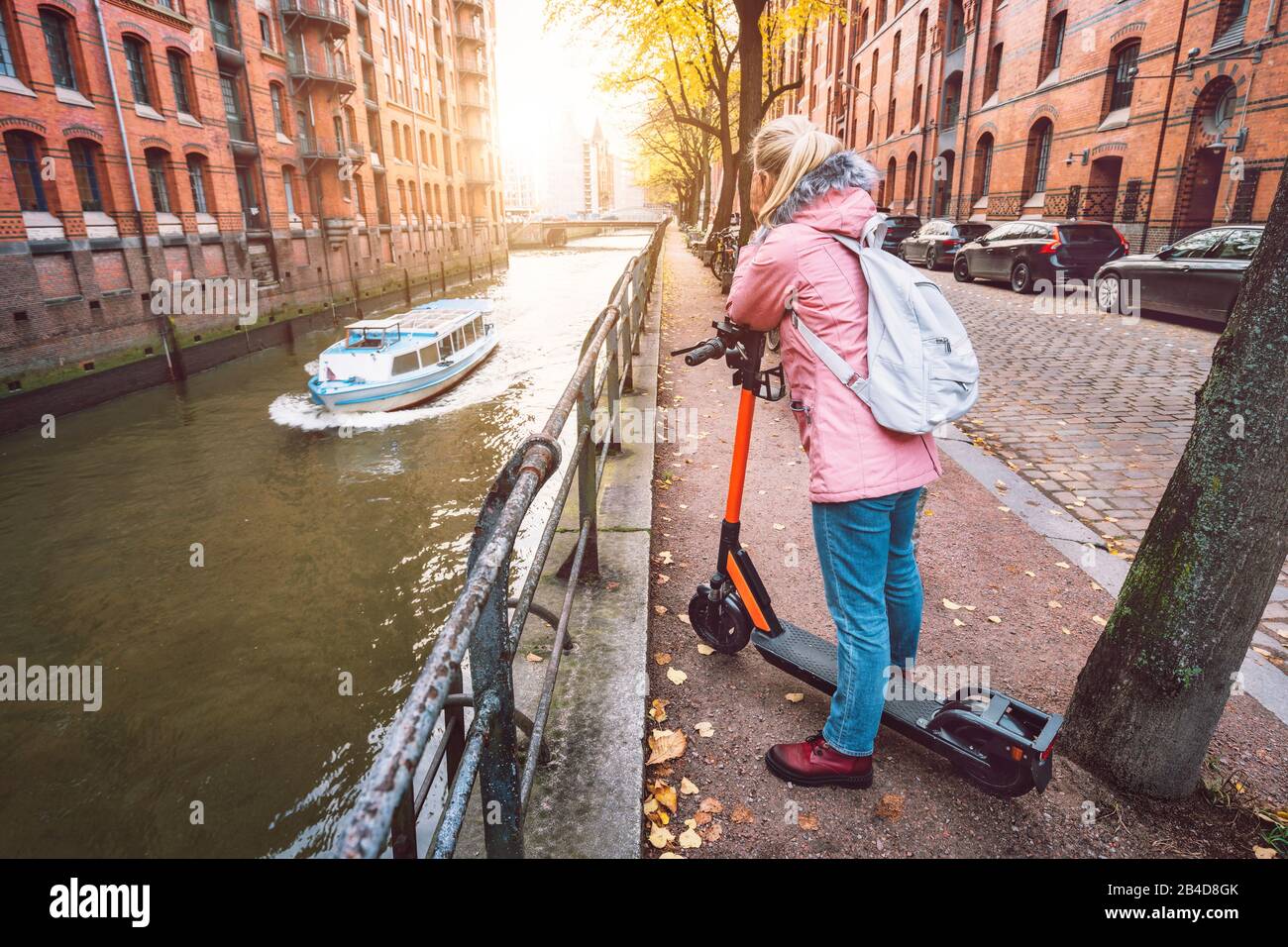 Rückenansicht einer Frau mit Rucksack und E-Roller, Blick auf die historische Speicherstadt in Hamburg im warmen Licht des Sonnenuntergangs, Deutschla Stock Photo