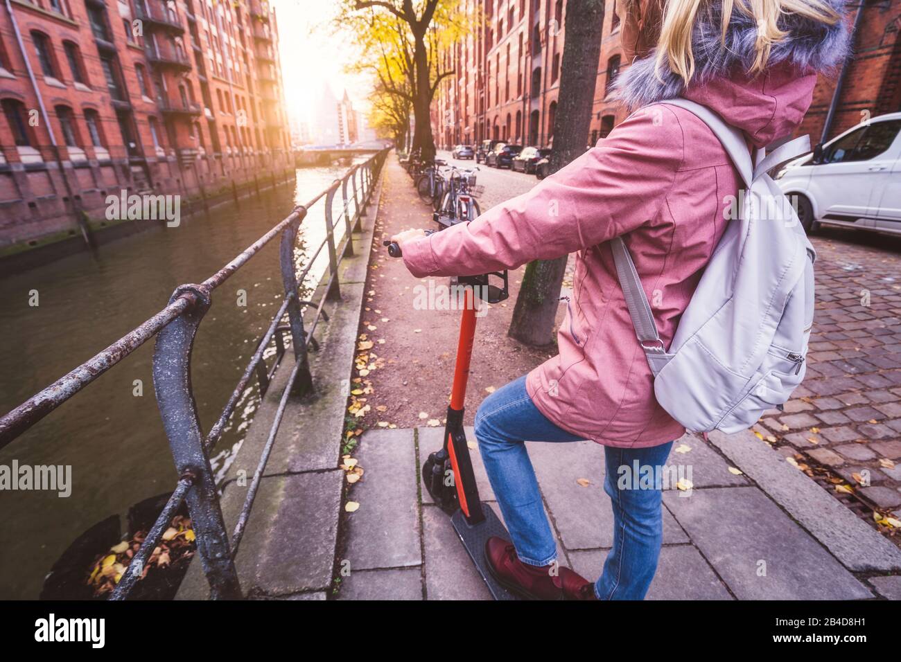 Rückenansicht einer Frau mit Rucksack und E-Roller, Blick auf die historische Speicherstadt in Hamburg im warmen Licht des Sonnenuntergangs, Deutschla Stock Photo
