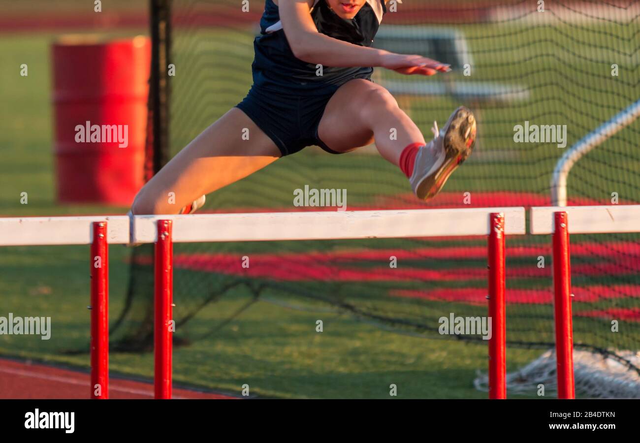 A high school girl is running the hurdles during a track and field competition outdoors on a sunny afternoon Stock Photo