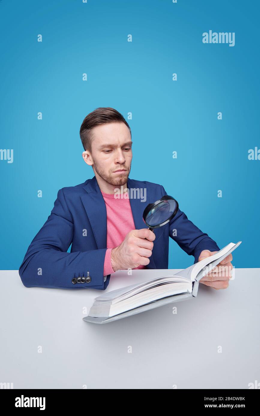Serious male reader with magnifying glass sitting by desk and looking through text on page of book on blue background Stock Photo