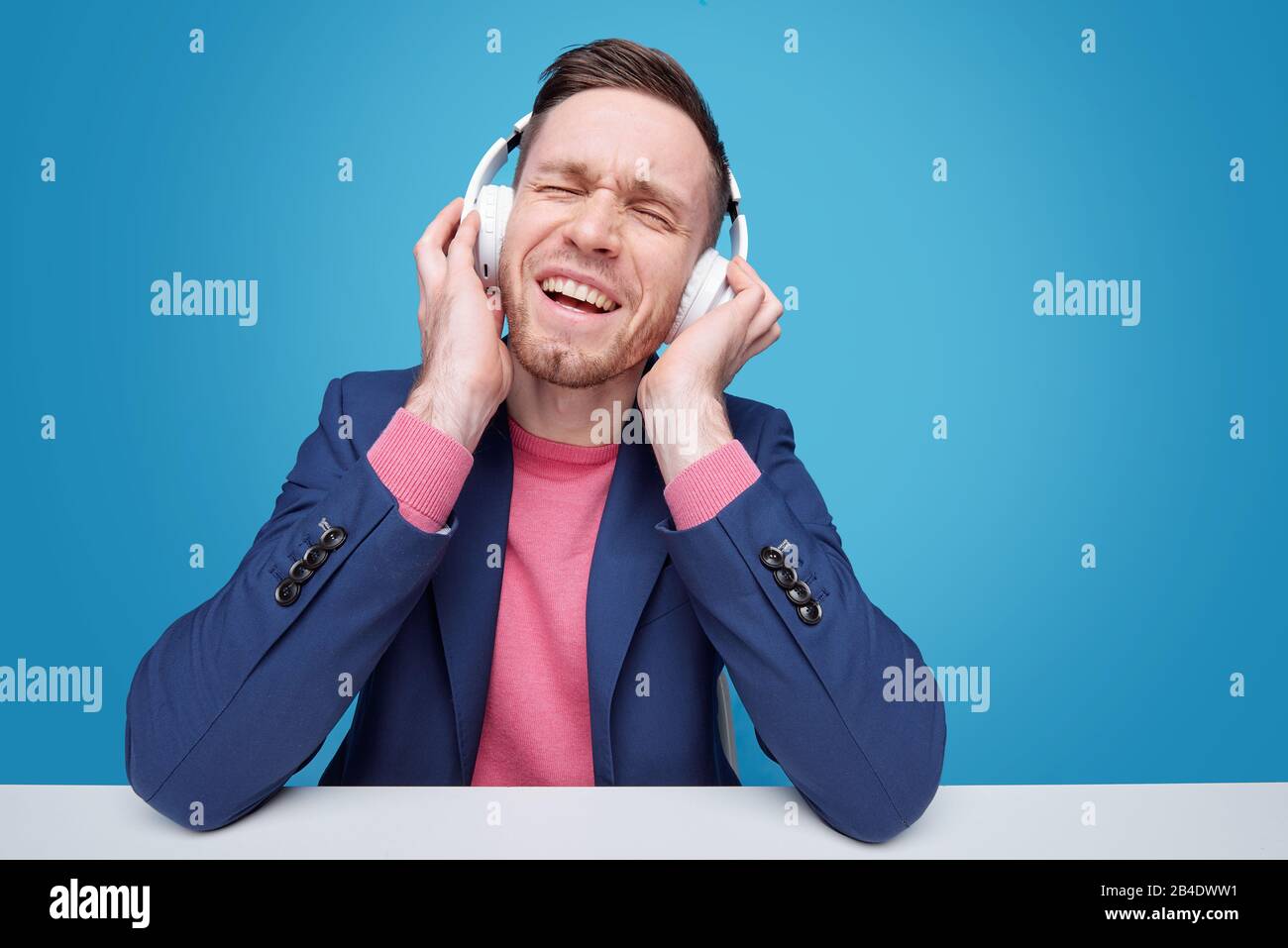 Cheerful and excited young man with headphones enjoying his favrite music while sitting by desk on blue background Stock Photo