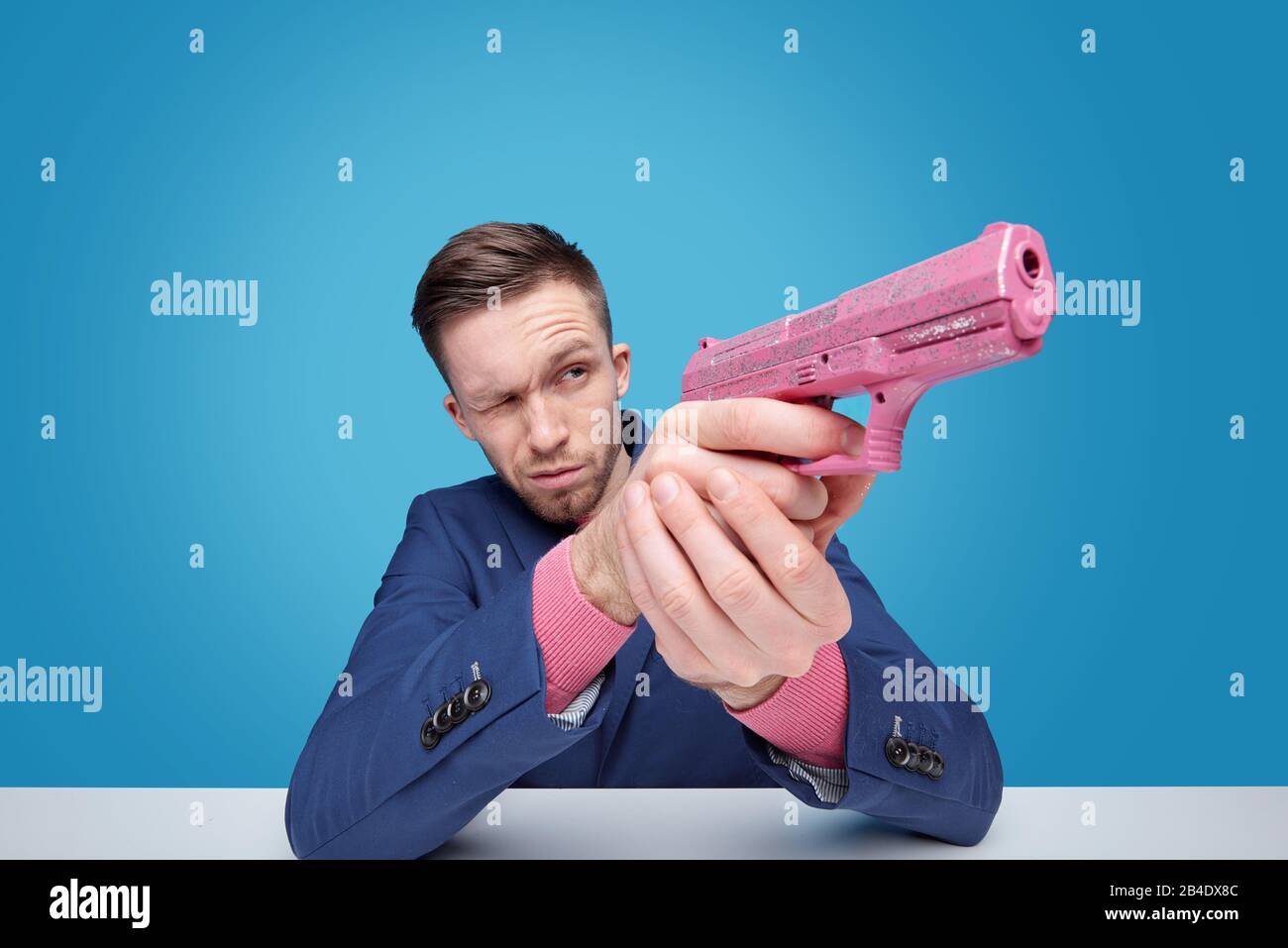 Young man with pink plastic toy gunaiming aside while sitting by desk in isolation over blue background Stock Photo