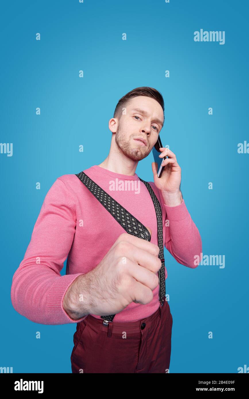 Confident young businessman stretching suspender while standing in front of camera and communicating on smartphone Stock Photo